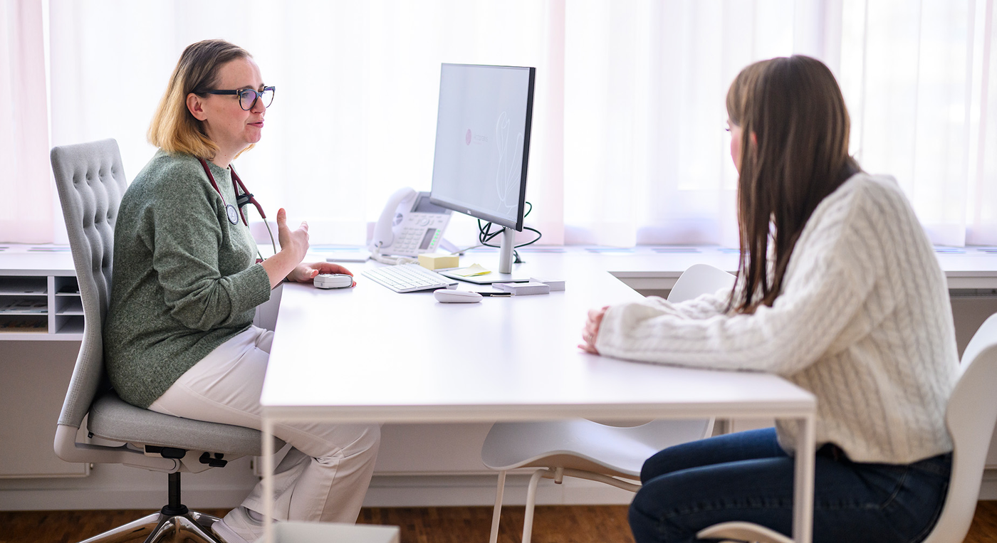 Aerztin sitzt im Behandlungszimmer am Schreibtisch vor dem Bildschirm und spricht mit einer Patientin